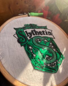 Hand-embroidered Slytherin house crest in green thread on fabric, displayed in an embroidery hoop